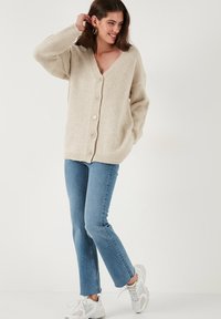 Cardigan beige lavorato a maglia con scollo a V, chiusura a bottoni e orlo a costine. Indossato sopra jeans blu, abbinato a sneakers bianche.