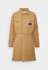 Veste en jean longue couleur beige clair, boutonnée, avec col, poches poitrine, poches latérales et petit écusson noir sur la poitrine gauche.
