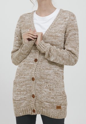 Femme portant un cardigan beige et blanc en maille avec des boutons et deux poches devant sur une chemise blanche, les mains jointes près de la poitrine.