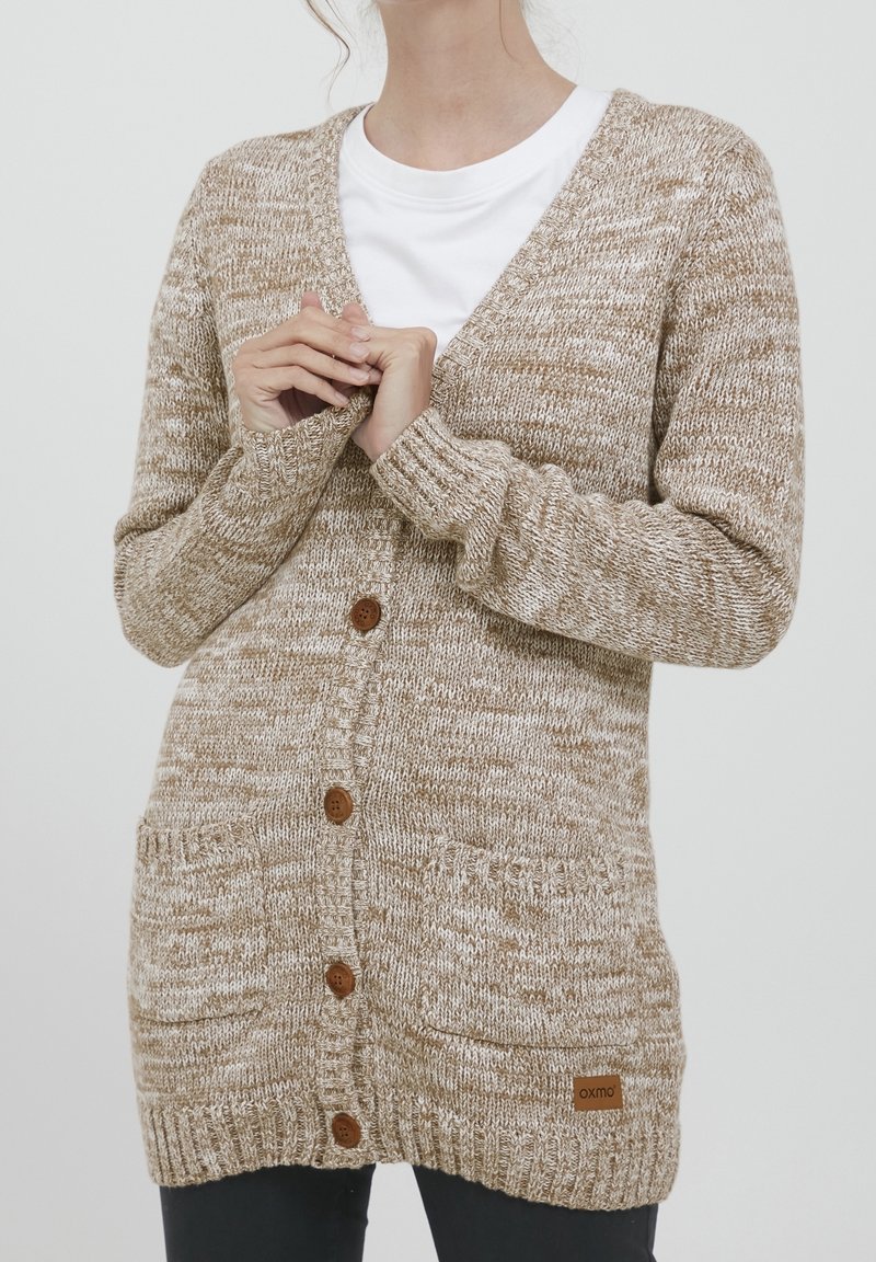 Femme portant un cardigan beige et blanc en maille avec des boutons et deux poches devant sur une chemise blanche, les mains jointes près de la poitrine.