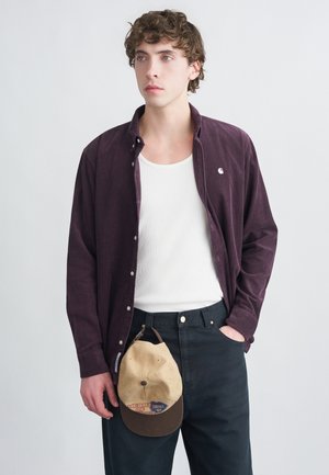 Dunkellila Cordhemd, weißes Tanktop darunter, schwarze Hose und eine beigefarbene Baseballkappe mit braunem Schirm, die aus der Tasche hängt.