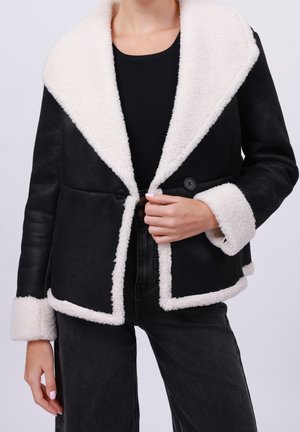 Vrouw draagt een zwart jack met een witte schapenvachtkraag, manchetten en rand, gecombineerd met een zwarte broek en een zwart topje.