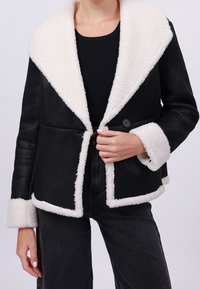 Femme portant une veste noire avec col, poignets et bordure en peau lainée blanche, associée à un pantalon noir et un haut noir.