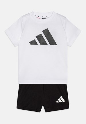 Tricou alb cu mâneci scurte pentru copii mici, cu un logo mare Adidas negru, asortat cu pantaloni scurți negri, cu un logo Adidas alb, mai mic.