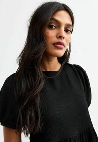 Abito nero con texture e maniche a sbuffo corte, caratterizzato da un design a strati e da un motivo sottile. Capelli scuri che scendono sulla spalla.
