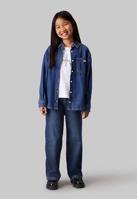 Chemise en jean boutonnée à manches longues, t-shirt blanc à motif en dessous, et jean en denim à jambes larges. Des chaussures noires complètent la tenue.