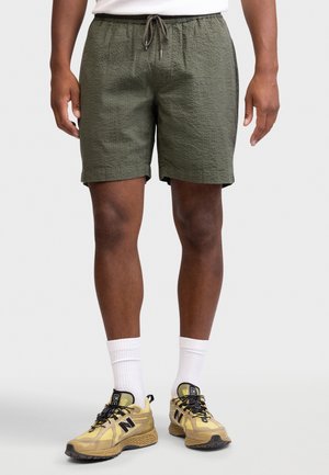 Mand iført olivengrønne, teksturerede shorts med snøre, hvide crew-sokker og gule New Balance-sneakers, stående foran en ensfarvet baggrund.
