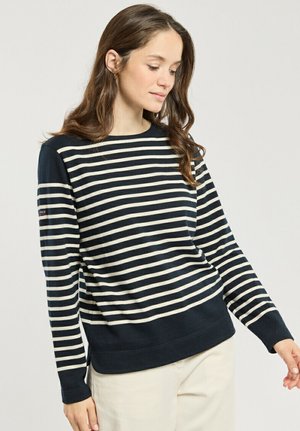 Femme portant un haut à manches longues rayé horizontalement bleu marine et blanc avec poignets et ourlet bleu marine, associé à un pantalon clair, debout devant un fond uni.