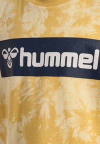 Gul t-shirt med ett strukturerat, blekt mönster; har en marinblå rand som visar märket "hummel" i vita blockbokstäver.