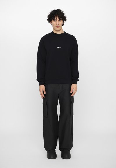 Sweatshirt noir avec des poignets et un ourlet côtelés, arborant un petit logo blanc. Associé à un pantalon cargo noir ample et des chaussures noirs à enfiler.