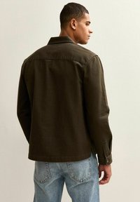 Chaqueta de mezclilla verde oliva con cuello, puños abotonados y detalles en el yugo trasero, combinada con jeans de color azul claro. Diseño simple y estructurado.