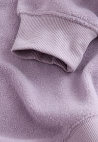 Close-up af en lavendel fleeceærme med ribstrikket manchet på en tekstureret, blød stofoverflade.