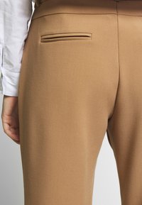 Beiga skräddarsydda byxor med en slät yta, som har en diskret bakficka, placerad nära midjebandet.