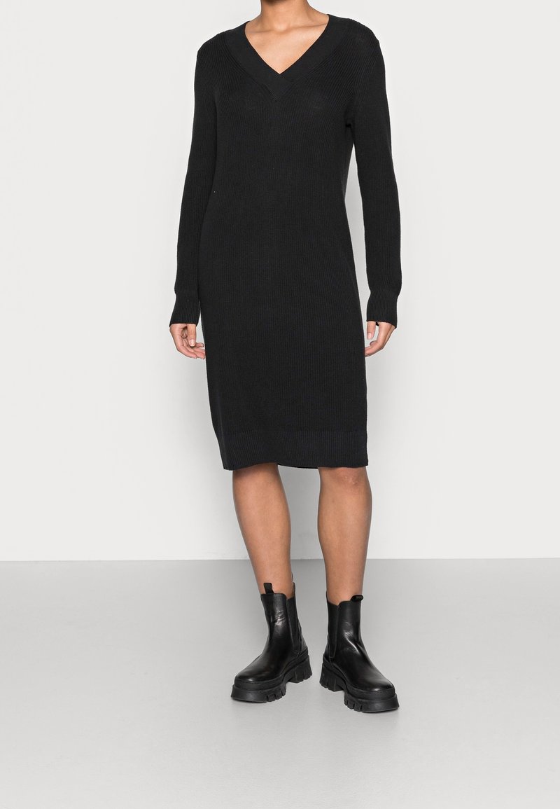 Femme portant une robe noire à manches longues en maille côtelée avec un col en V, associée à des bottines en cuir noir épaisses, debout sur un sol gris clair.