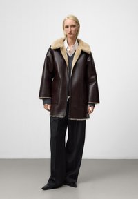 Cappotto in pelle marrone con colletto in pelliccia, dotato di zip frontale, grandi tasche e una vestibilità rilassata. Indossato sopra strati grigi e bianchi.