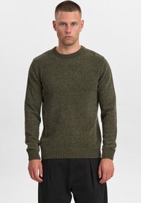 Maglione lavorato a maglia verde oliva con scollo rotondo, polsini e orlo a coste; tessuto strutturato con un motivo sottile, indossato con pantaloni neri.