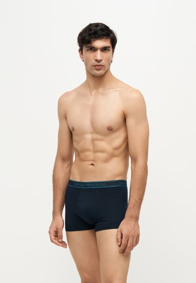 Jeune homme aux cheveux courts et foncés portant un boxer noir Emporio Armani, debout devant un fond clair uni.