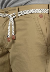 Pantaloni kaki cu o suprafață texturată, o curea de tip frânghie în alb cu accente bleumarin și un patch din piele maro cu logo pe talie.