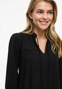 Melnas garām piedurknēm blūze ar V-veida izgriezumu, kurai ir smalks saišu akcents ap kaklu un viegli pūkaini pleci. Maiga auduma tekstūra.