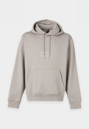 Junger Mann mit geflochtenem Haar, der einen grauen Hoodie, weit geschnittene hellgraue Jeans und sportliche Sneaker trägt, vor einem schlichten weißen Hintergrund stehend.