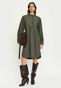 Robe verte à manches longues avec col à volants, détails de boutons et corsage évasé. Associée à un sac en suède marron et des bottes marron jusqu'aux genoux.