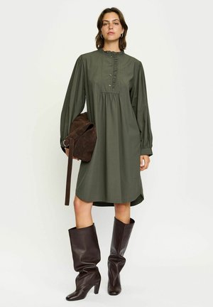 Groene jurk met lange mouwen, rimpelcol en knoopdetails, met een uitlopende taille. GeCombineerd met een bruine suède tas en kniehoog bruine laarzen.