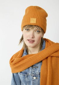 Orange knit beanie with a ribbed texture and square logo patch, paired with an orange knit scarf, draped around shoulders over a denim jacket.