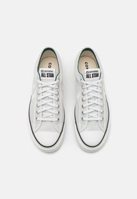 Vita kanvas sneakers med låg design, som har en gummitå, svart rand och gröna accenter på insidan och märket.