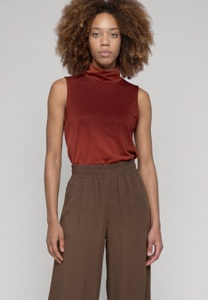 Vrouw met krullend haar die een mouwloos roestkleurig coltopje en loszittende bruine broek draagt, staand tegen een effen achtergrond.