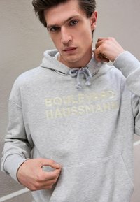 Gray hoodie featuring "BOULEVARD HAUSSMANN" in light yellow text, drawstring neck, and front pocket. Soft fabric with ribbed cuffs.
