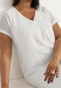 Kvinna som bär en kortärmad vit spetsblus med V-ringning, sitter med en arm vilande, bär en dubbel silverring och mörk nagellack.