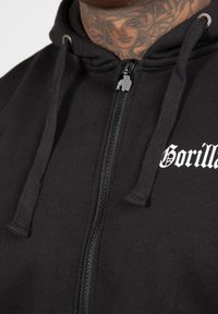 Melnā, uzvelkama kapuce, izgatavota no mīkstas auduma, ar priekšējo rāvējslēdzēju, kuram ir gorillas formas vilkšanas elements, un ar baltu "Gorilla" tekstu uz krūšu.