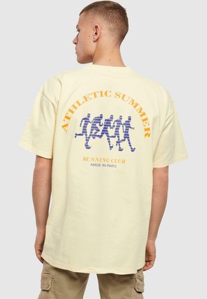 T-shirt overdimensionato color crema con una grafica di figure in corsa accompagnata da testi blu e arancioni: "CLUB DI CORSA ESTIVO ATLETICO MADE IN PARIS."
