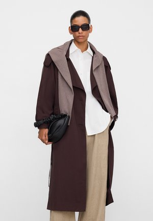 Vrouw draagt een oversized donkerbruine jas, wit overhemd, beige broek, grijze sjaal over de schouders gedrapeerd, zwarte zonnebril en houdt een zwarte geplooide clutch vast.