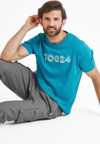 Turquoise cotton t-shirt with "TOG24" logo in white and yellow. Gray cargo pants with pockets. Model sitting, relaxed pose.