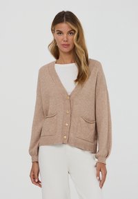 Donna in cardigan beige con bottoni e tasche frontali, indossato sopra una maglietta bianca e pantaloni bianchi, in piedi contro uno sfondo chiaro e uniforme.