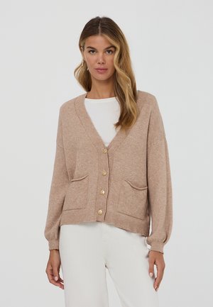 Donna in cardigan beige con bottoni e tasche frontali, indossato sopra una maglietta bianca e pantaloni bianchi, in piedi contro uno sfondo chiaro e uniforme.
