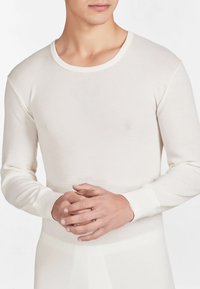 Maglione termico bianco a maniche lunghe con texture a coste, scollatura tonda e polsini aderenti. Il materiale sembra morbido e leggero. Mani incrociate davanti.