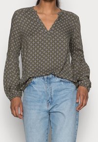 Blouse vert olive avec un col en V, manches longues bouffantes et motifs floraux blancs. Associée à un jean taille haute bleu clair.