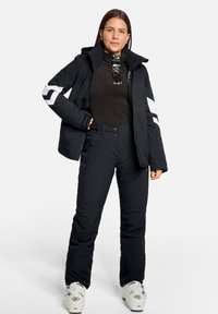 Schwarze isolierte Skijacke mit kontrastierenden weißen Streifen und durchgehendem Reißverschluss, kombiniert mit passenden schwarzen Hosen und Skischuhen.