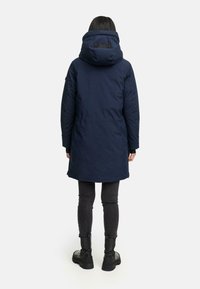 Person står med ryggen til iført en marineblå hoodie-parka, tætsiddende mørke jeans og sorte kraftige støvler på en hvid baggrund.