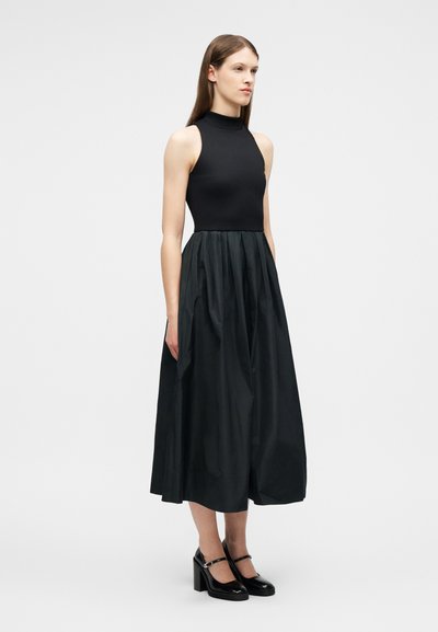 Femme portant un haut noir sans manches à col haut et une jupe midi noire volumineuse avec des chaussures Mary Jane noires, debout devant un fond blanc uni.