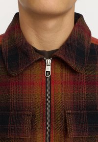 Geruite jas met een mix van rode, bruine en zwarte kleuren, met een ritssluiting en een kraag. Gemaakt van een textuurachtig wolachtig materiaal.