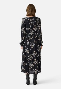 Black floral maxi dress with long sleeves, cinched waist, and flowing fabric. Features a leaf pattern in muted colors and button closure at the back.