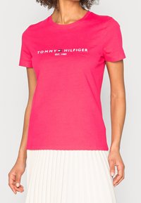 T-shirt a maniche corte in cotone rosa brillante con logo bianco "TOMMY HILFIGER" e testo "EST. 1985", scollatura rotonda e vestibilità rilassata.