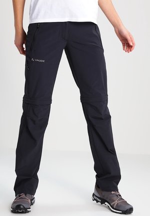 Schwarze Wanderhose mit Reißverschlussbeinen und Seitentaschen, getragen mit grauen und pinken Wanderschuhen vor weißem Hintergrund.