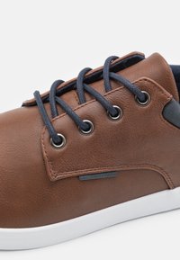 Sneaker en synthétique marron avec un design à lacets bleu marine, œillets en métal argenté, semelle en caoutchouc blanche et un panneau d'accent noir.