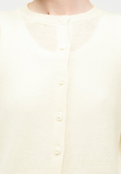 Κοντινή λήψη κρεμ πλεκτής ζακέτας με στρογγυλή λαιμόκοψη, εμπρόσθιο κούμπωμα και διακοσμητικό δίχτυ στο ανώτερο μέρος του στήθους.