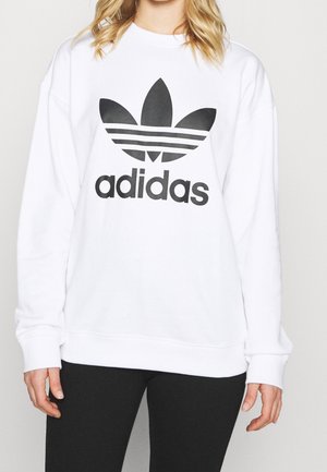 Personne portant un sweat Adidas blanc avec un grand logo trefoil noir et un texte, associé à un pantalon noir, sur un fond uni.