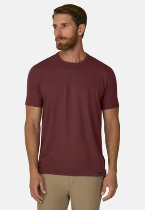 Uomo con la barba che indossa una maglietta a maniche corte bordeaux uniforme e pantaloni beige, in piedi davanti a uno sfondo chiaro e uniforme.